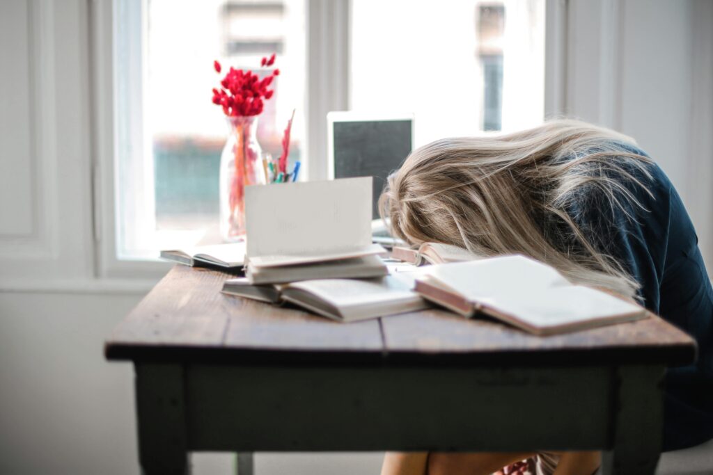 pexels-photo-3767411-3767411 Blonde woman slumped over desk filled with books, showcasing study fatigue indoors.
